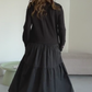 O vestido apresenta um decote com fecho, mangas compridas e uma saia plissada em camadas.